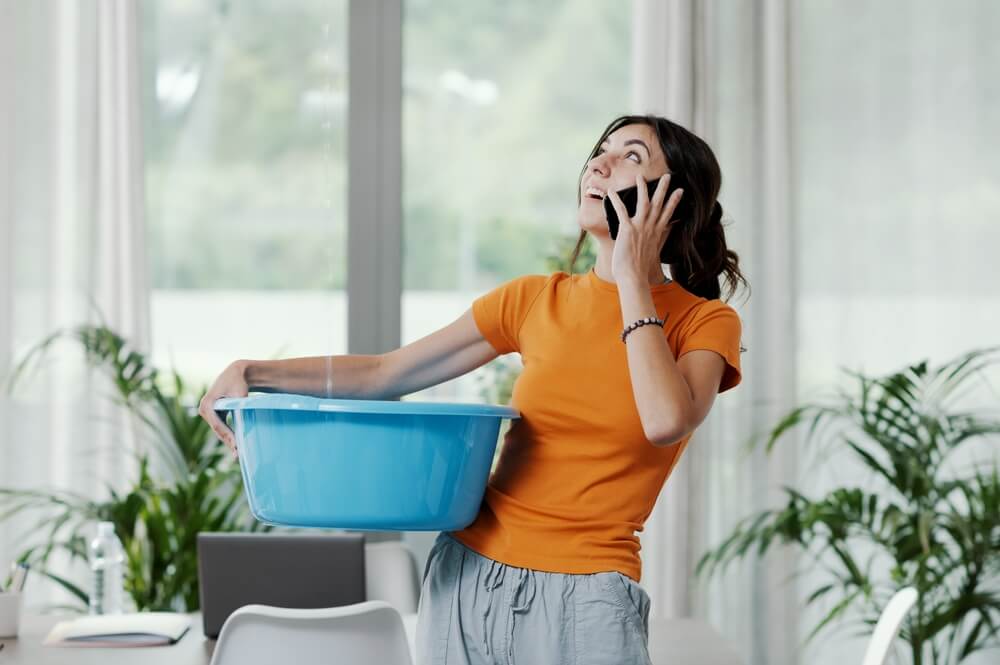 A woman holding a tub to catch water leaking from the ceiling while on the phone